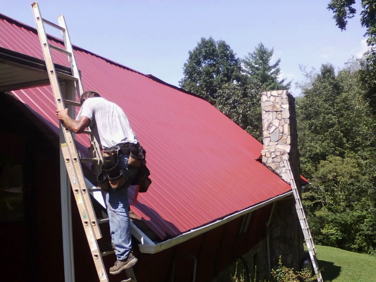 Roofer on a ladder beside a red metal roof and stone chimney — Soffit & Fascia Repair by Roofing Brothers in Pittsfield